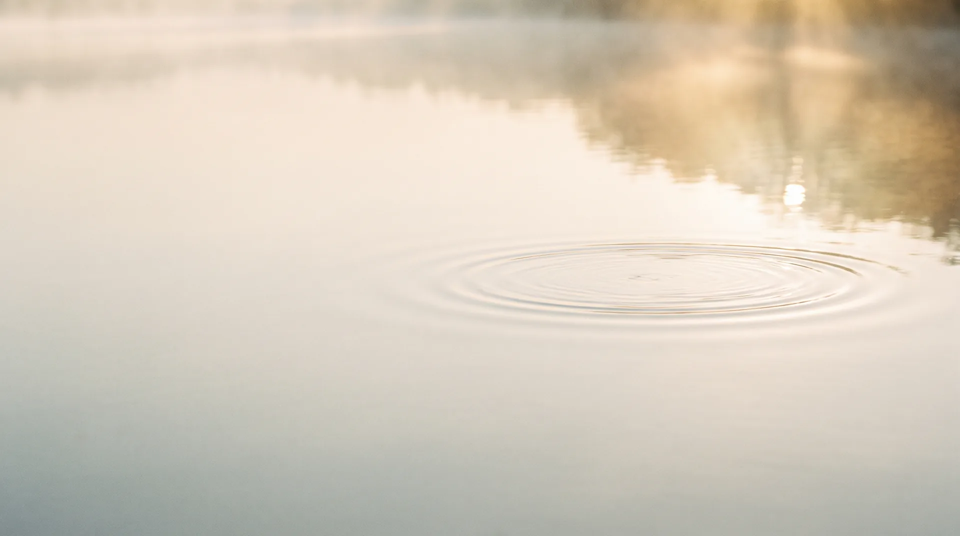 Still water at dawn with a single ripple spreading in soft golden light