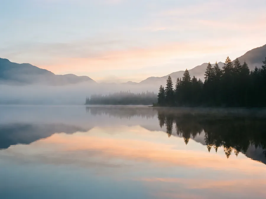 A calm, misty lake at sunrise reflecting a soft pastel sky